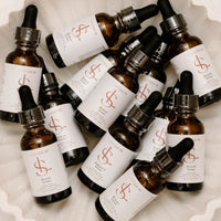 A group of amber glass dropper bottles with white labels and black caps, arranged in a white scalloped dish. The labels feature a prominent coral-red dollar sign logo and the brand name LUMISKIN.