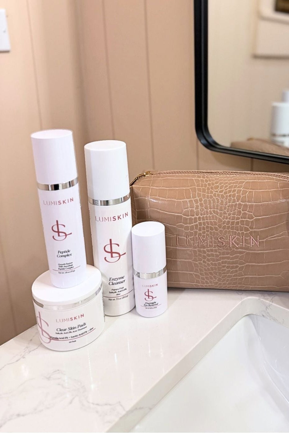 A set of white and silver skincare products from the brand LUMISKIN, including bottles, jars, and a textured tan zippered pouch, arranged on a bathroom counter next to a sink and mirror.