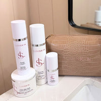 A set of white and silver skincare products from the brand LUMISKIN, including bottles, jars, and a textured tan zippered pouch, arranged on a bathroom counter next to a sink and mirror.