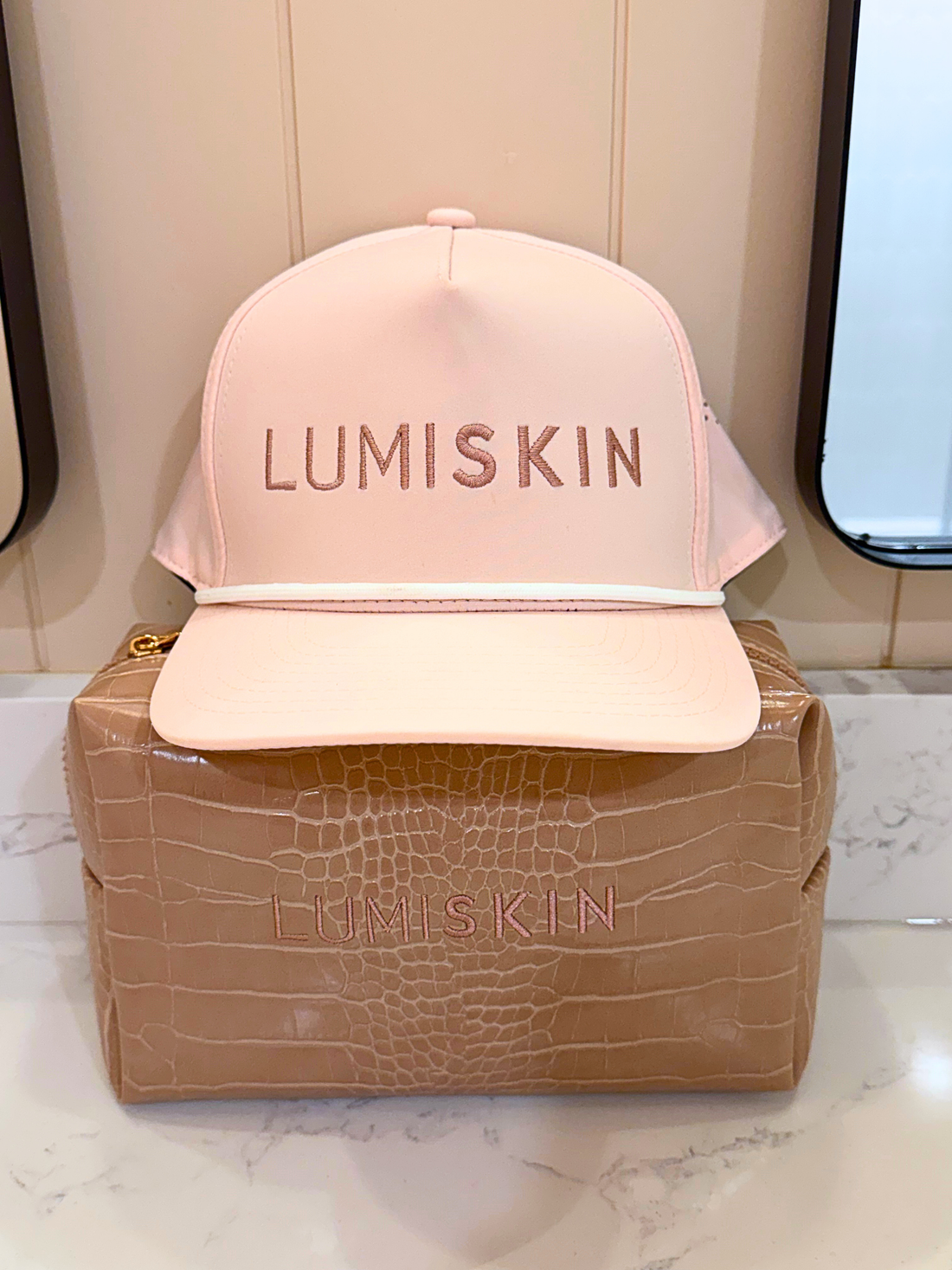 A pale pink LUMISKIN baseball cap with tonal embroidered lettering, displayed on top of a tan crocodile-embossed pouch. The background shows a beige paneled wall and a mirror.