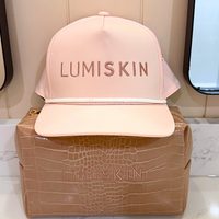 A pale pink LUMISKIN baseball cap with tonal embroidered lettering, displayed on top of a tan crocodile-embossed pouch. The background shows a beige paneled wall and a mirror.