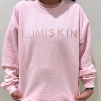 A person wearing a light pink crewneck sweatshirt with 'LUMISKIN' printed across the chest in a matching pink tone, shown against a plain light gray wall.