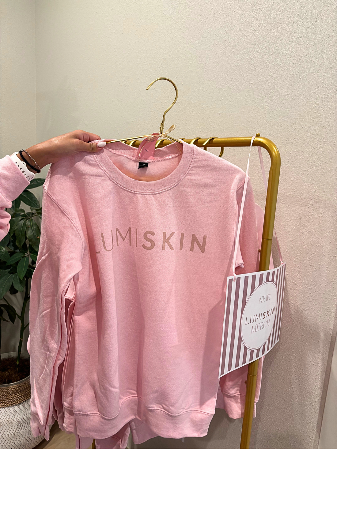 A pink crewneck sweatshirt with 'LUMISKIN' printed across the chest in a metallic rose-gold tone, held up by a hand on a gold hanger. The sweatshirt is displayed on a gold clothing rack next to a striped paper tag.