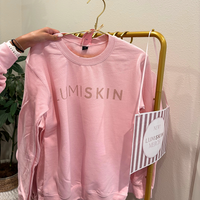 A pink crewneck sweatshirt with 'LUMISKIN' printed across the chest in a metallic rose-gold tone, held up by a hand on a gold hanger. The sweatshirt is displayed on a gold clothing rack next to a striped paper tag.