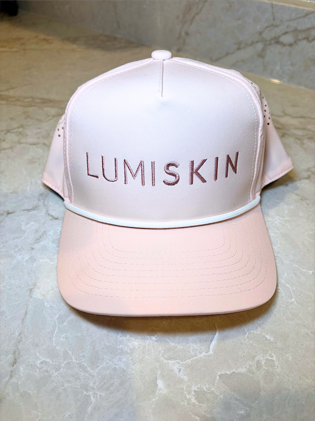 A light pink trucker-style cap with 'LUMISKIN' embroidered in rose gold on the front, resting on a beige marble surface.