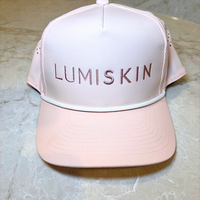 A light pink trucker-style cap with 'LUMISKIN' embroidered in rose gold on the front, resting on a beige marble surface.