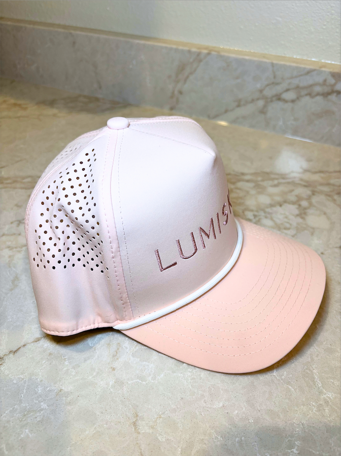 A light pink baseball cap with a matching curved brim, featuring a perforated mesh back panel and the brand name 'LUMISKIN'.