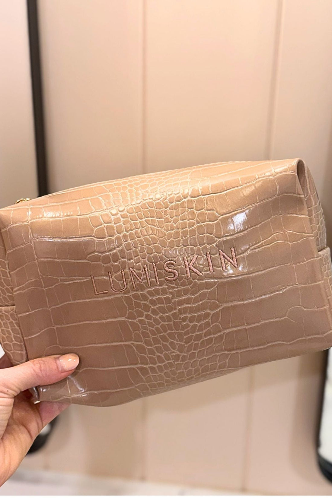 A hand holding a beige crocodile-embossed cosmetic pouch with the brand name 'LUMISKIN' debossed on the front, set against a neutral indoor background.