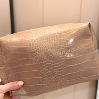 A hand holding a beige crocodile-embossed cosmetic pouch with the brand name 'LUMISKIN' debossed on the front, set against a neutral indoor background.