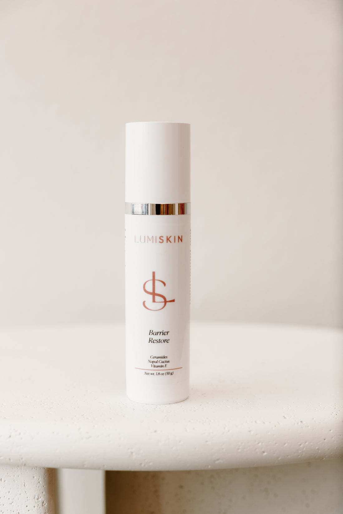 A white cylindrical skincare bottle with a metallic silver band and a mauve colored logo, standing on a textured white surface against a soft beige background. The brand name 'LUMISKIN' is visible on the bottle.
