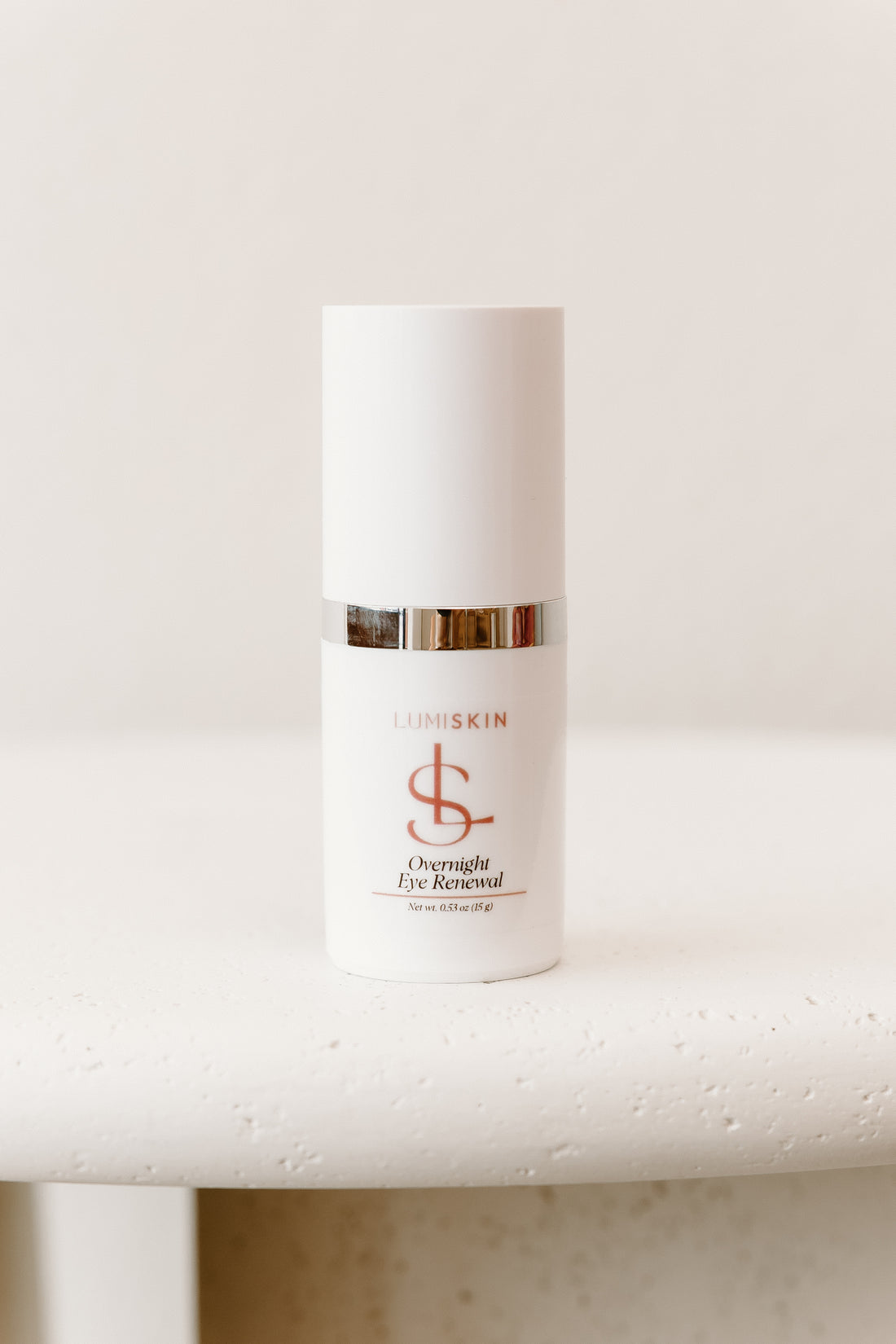 LUMISKIN Overnight Eye Renewal skincare bottle in white with a metallic gold band and mauve logo, resting on a textured white surface against a warm off-white background.