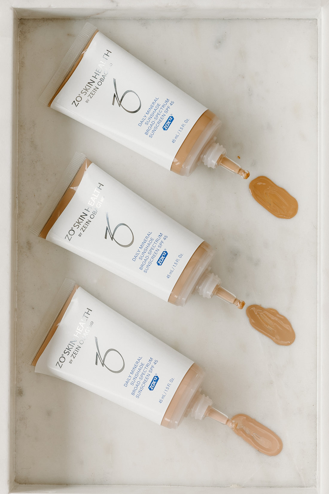 Three squeeze tubes of ZO SPF arranged diagonally on a white marble tray, with dollops of the beige foundation visible at the tips of the applicators.