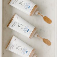 Three squeeze tubes of ZO SPF arranged diagonally on a white marble tray, with dollops of the beige foundation visible at the tips of the applicators.