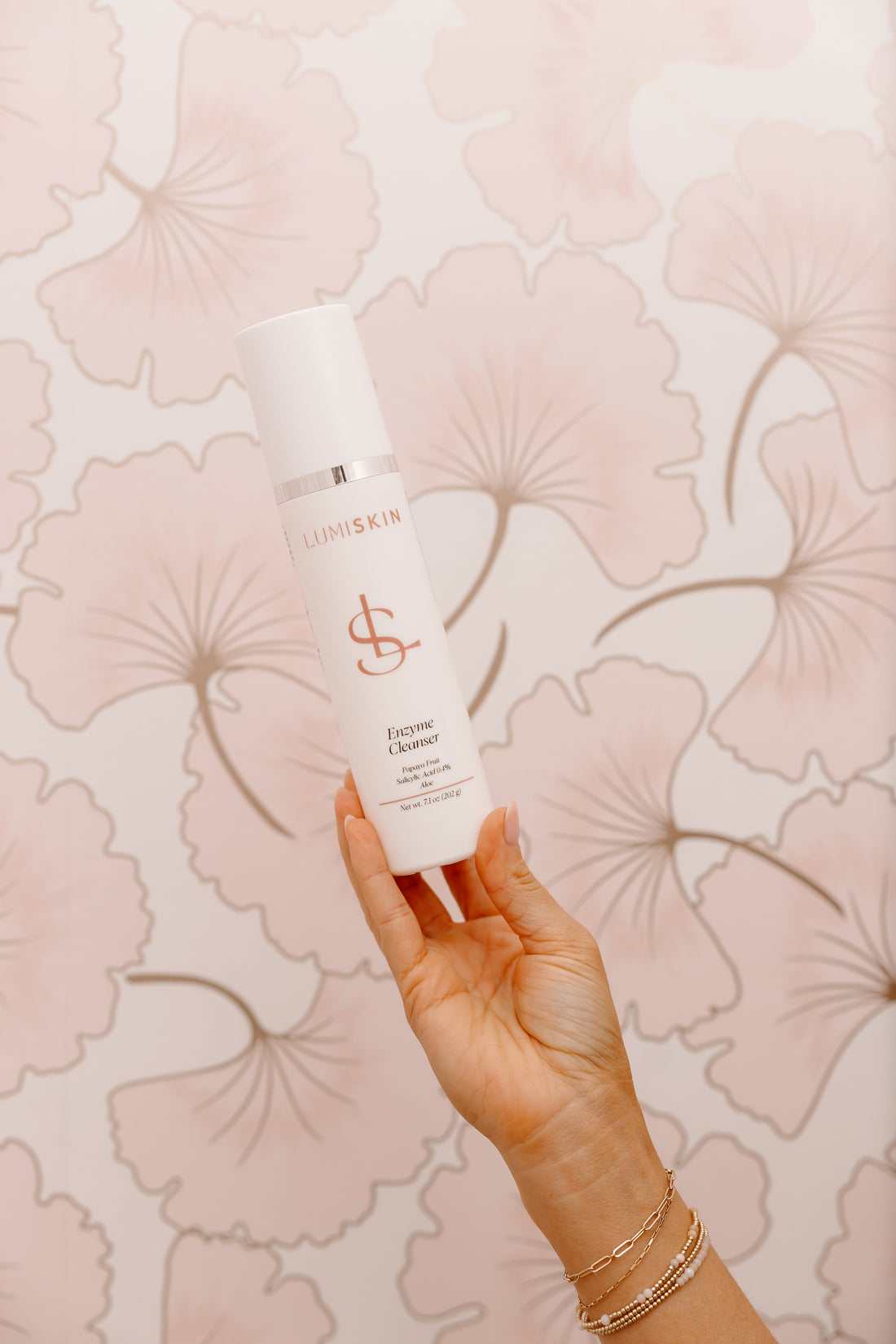 A hand holding a white LUMISKIN Enzyme Cleanser bottle against a pink floral wallpaper background.