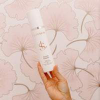 A hand holding a white LUMISKIN Enzyme Cleanser bottle against a pink floral wallpaper background.