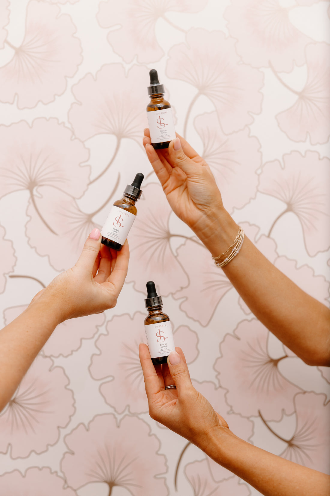 Three hands holding small amber dropper bottles with white labels against a pale pink floral patterned background.