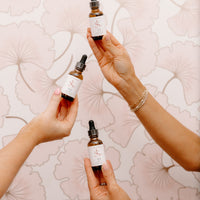 Three hands holding small amber dropper bottles with white labels against a pale pink floral patterned background.