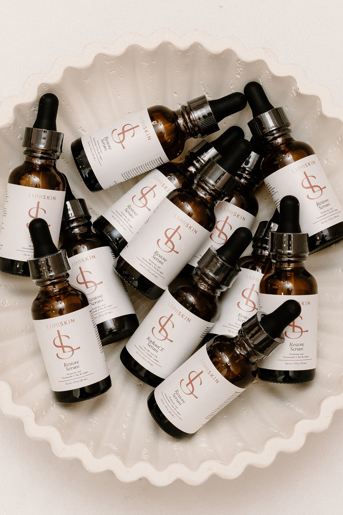 A group of amber glass dropper bottles with white labels and black caps, arranged in a white scalloped dish. The labels feature a prominent coral-red dollar sign logo and the brand name LUMISKIN.