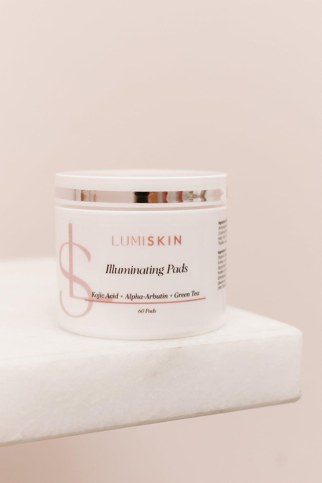 A white cosmetic jar labeled LUMISKIN Illuminating Puds, placed on a white marble-like pedestal against a soft blush pink background.